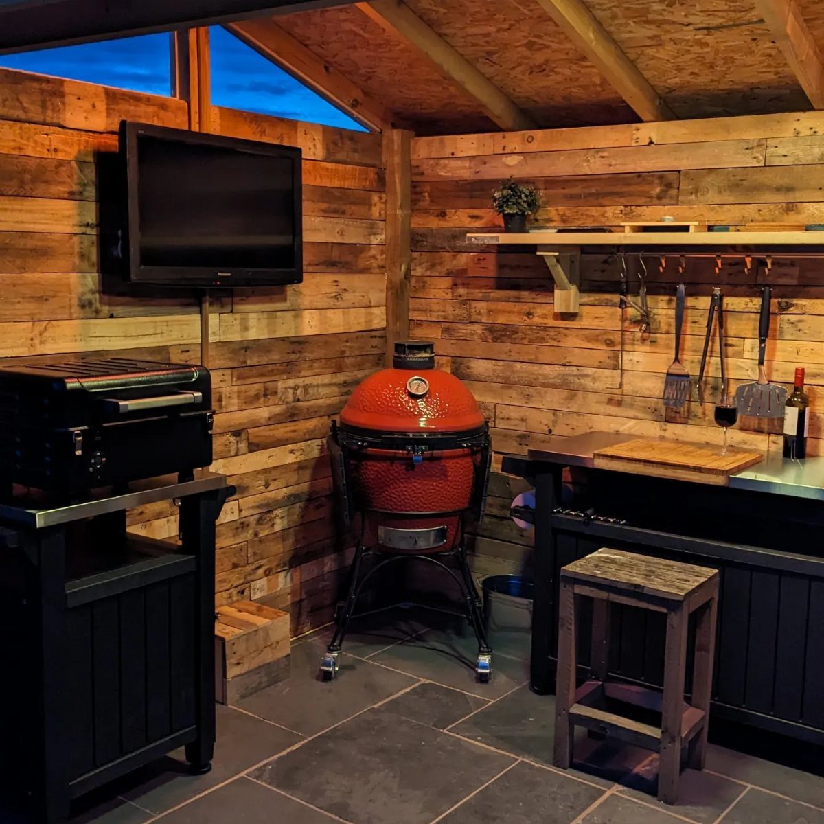 A rustic wooden outdoor kitchen shelter containing a red kamado grill, black gas grill, and a wall-mounted television.