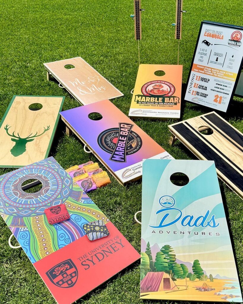Various custom-designed cornhole boards with colorful graphics resting on a green grass lawn.