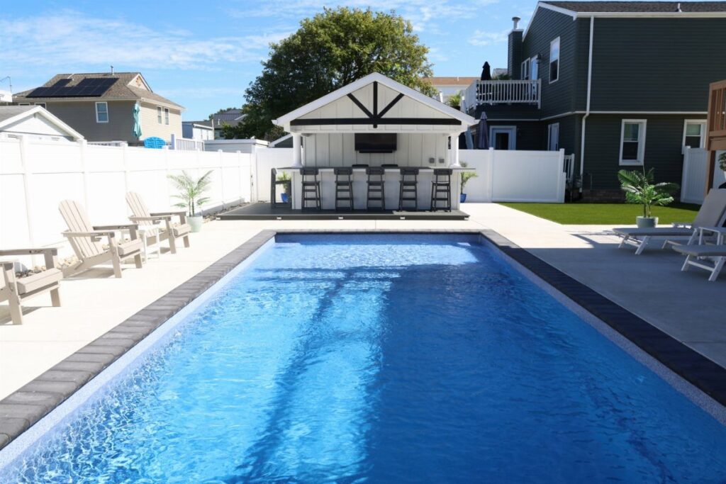 A rectangular blue swimming pool in a backyard next to a white poolside bar structure.