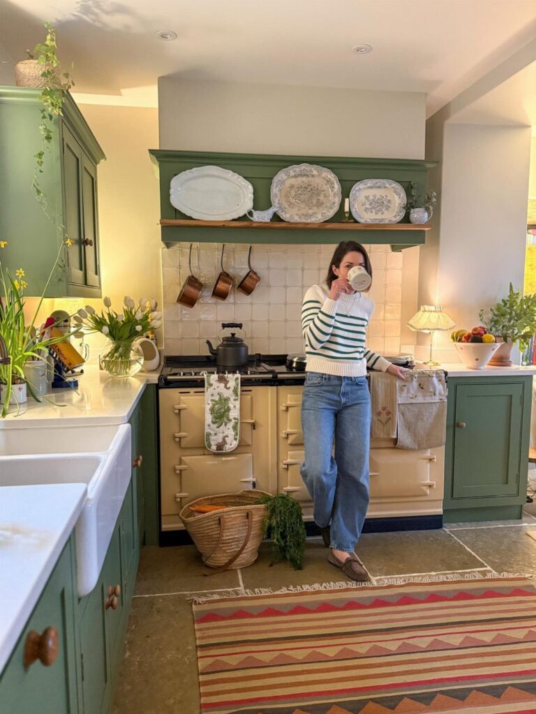 A woman sipping from a mug in a rustic sage green kitchen with an AGA stove.