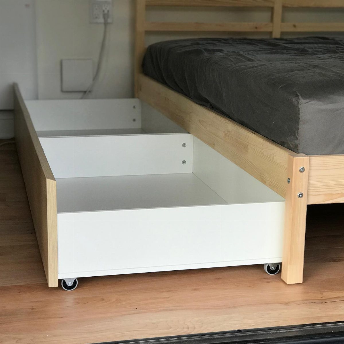 A white wooden storage drawer with wheels pulled out from under a light wood bed frame.