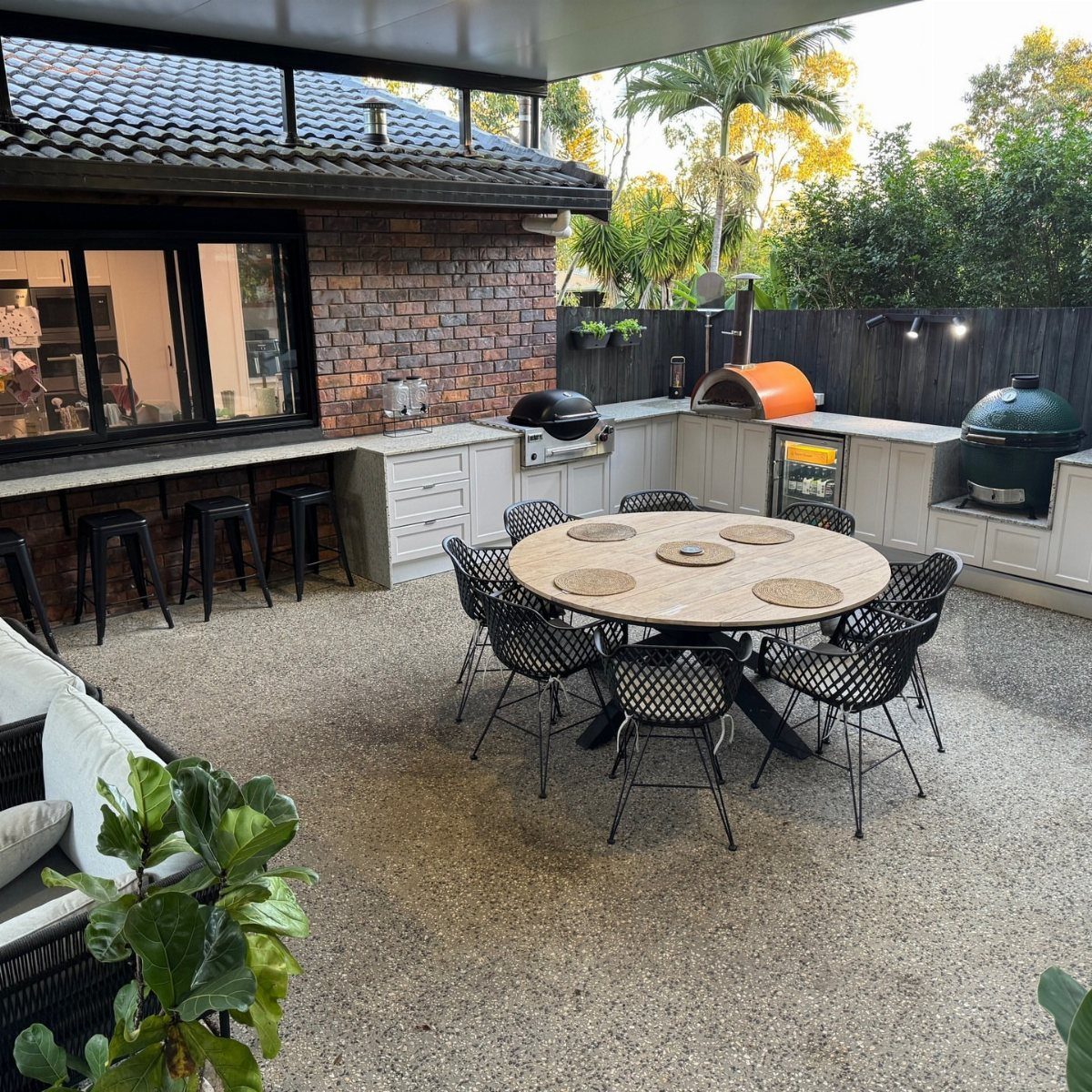 An L-shaped outdoor kitchen with grey cabinets and a grill set on a gravel patio with a dining table.