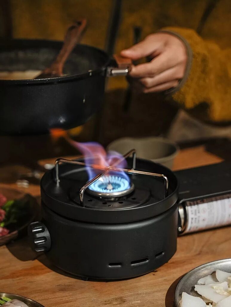 A portable black gas stove with a blue flame cooking food in a black pot.