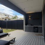 Grey composite decking boards leading to a modern dark outdoor kitchen under a covered patio.