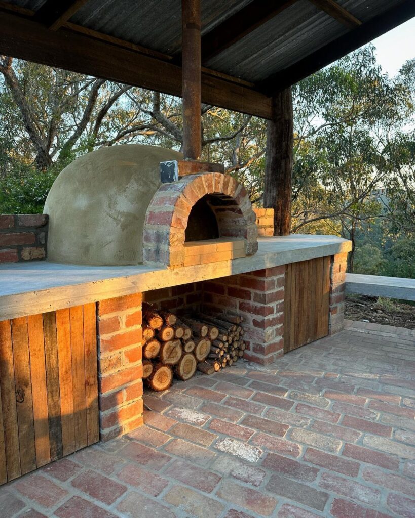 A brick outdoor pizza oven with a concrete countertop and wood storage under a wooden shelter.